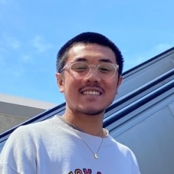 A Vietnamese person with a black buzzcut, clear glasses, and a bit of facial hair, standing against the vibrant blue sky.